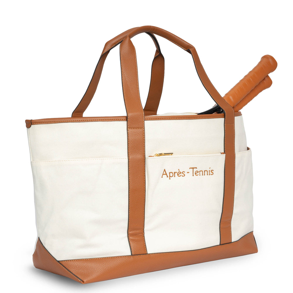 Back view of the Après‑Tennis Ultimate Tote in beige and brown, a structured canvas bag with vegan leather straps and gold hardware, shown with a tennis racquet inside and an embroidered brown “après-tennis” text on a pocket.