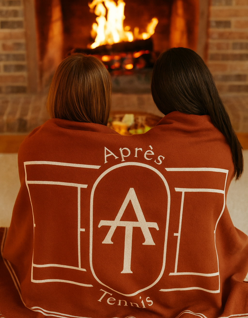 Two tennis-friends sitting by a fireplace, wrapped in a red clay coloured blanket with 'Apres Tennis' branding.